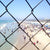  We are looking through a fence at a large crowd of people on the beach. In the background we see the mountains and blue skies. The photograph is printed on fine art paper.